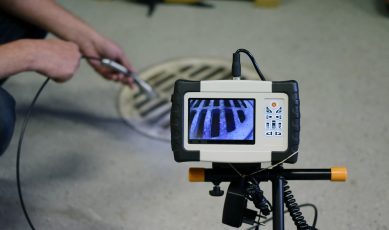 Un technicien d’Algora Environnement est en train de passer un câble dans une bouche d’égout afin de vérifier l’intérieur. Au premier plan on y voit une caméra d’inspection vidéo de canalisations affichant ce que le technicien vise.