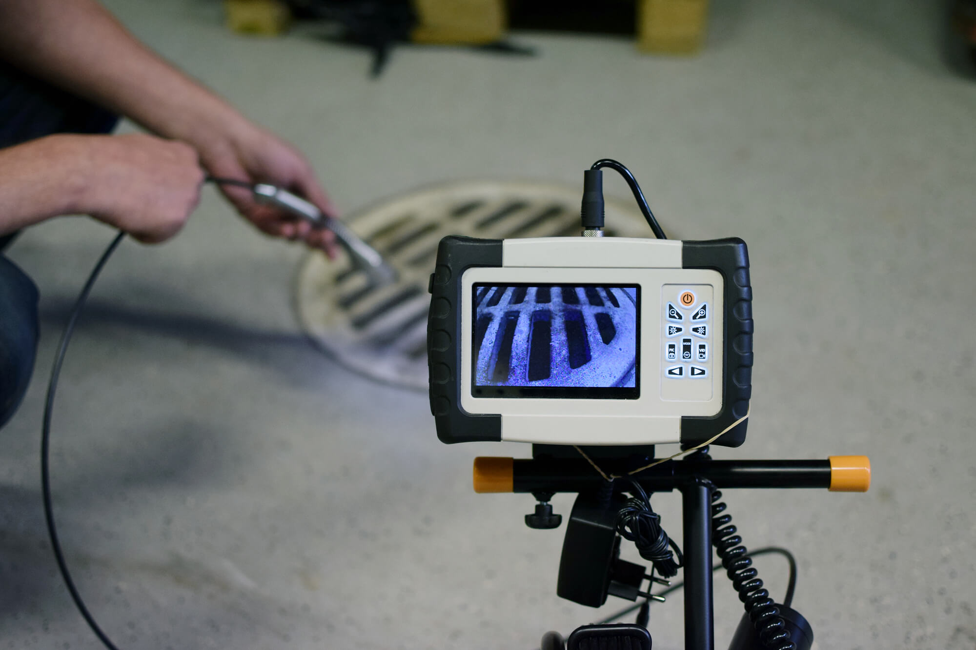Un technicien d’Algora Environnement est en train de passer un câble dans une bouche d’égout afin de vérifier l’intérieur. Au premier plan on y voit une caméra d’inspection vidéo de canalisations affichant ce que le technicien vise.